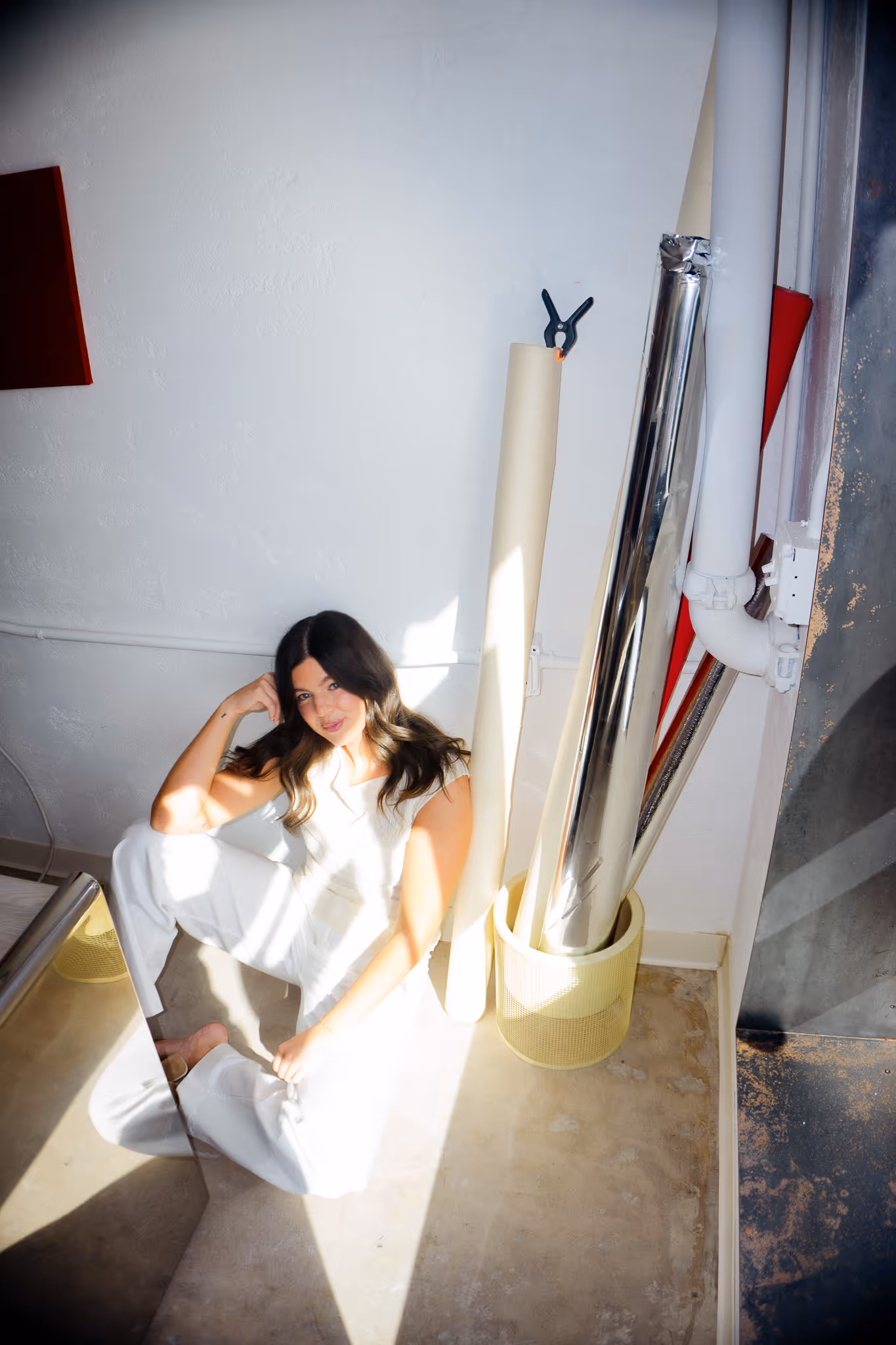 Woman with dark hair wearing white clothing sitting barefoot on a tiled floor beside rolled-up reflective and paper materials in a yellow basket near a white wall.