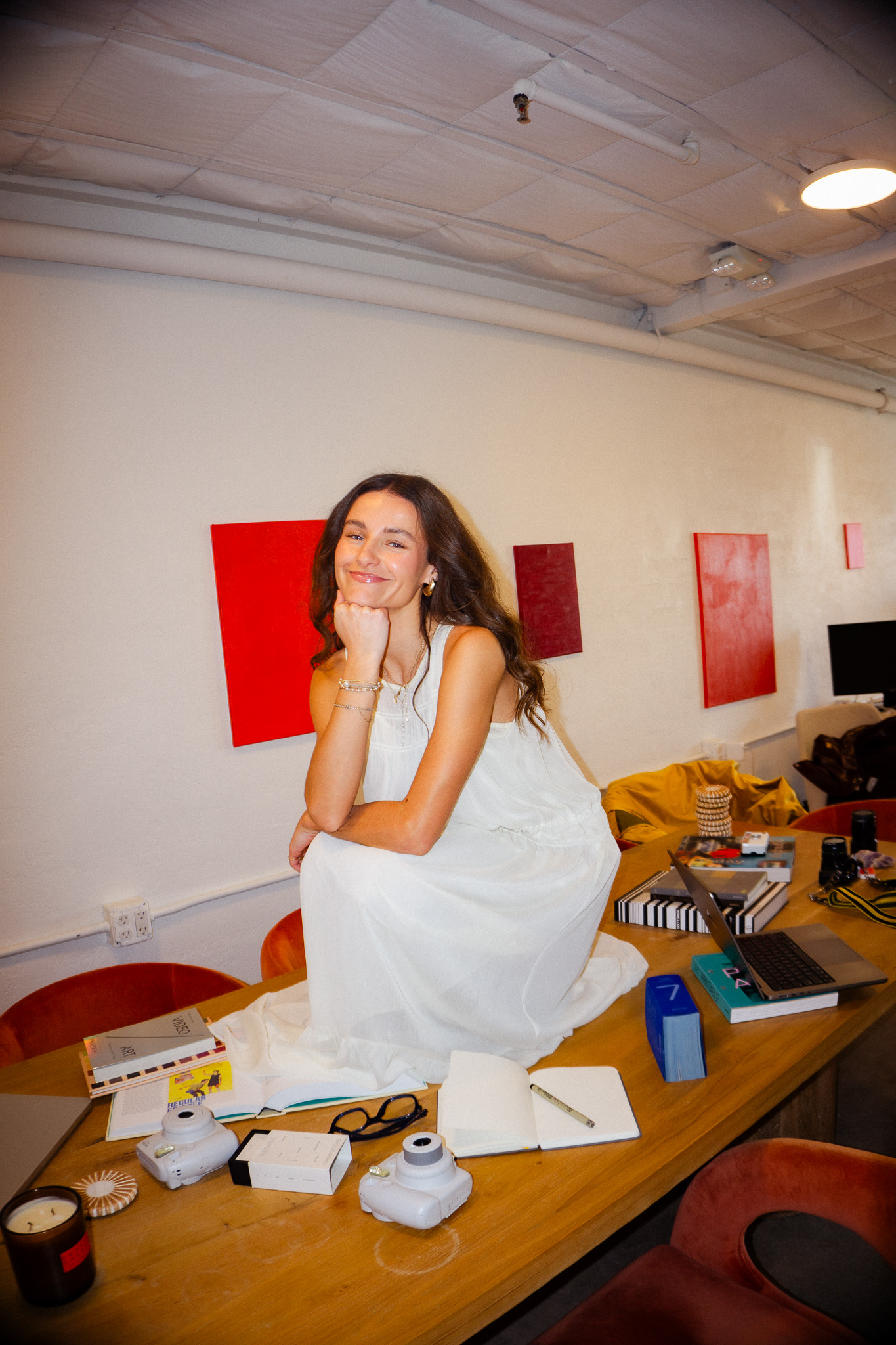 Smiling woman in a white dress sitting on a wooden table surrounded by books, a laptop, cameras, and other objects in a modern room with red wall art.