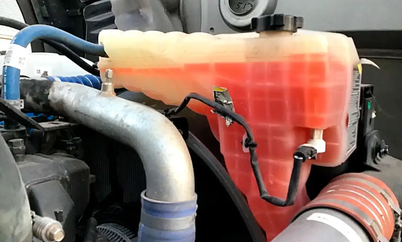 Close-up view of a diesel truck's translucent coolant reservoir filled with orange coolant, showing connected hoses and sensor wiring, located in the engine bay.