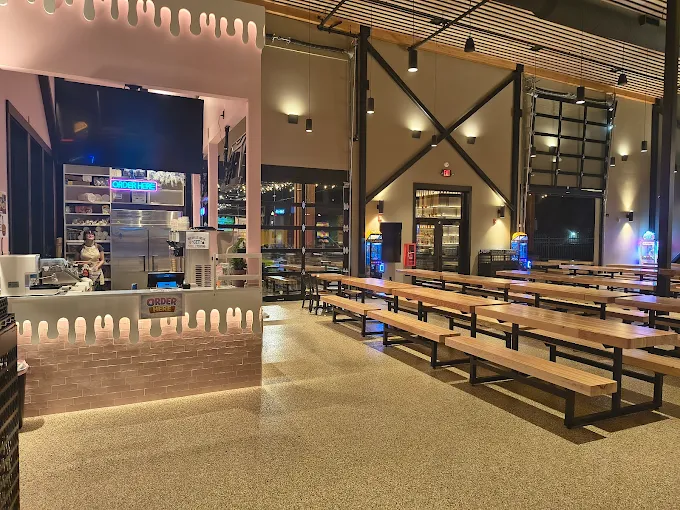 Interior of a spacious modern cafeteria with long wooden picnic-style tables and benches, a counter with 'Order Here' signs, and warm ambient lighting.