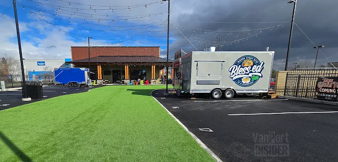 Outdoor area with artificial grass, string lights overhead, a building with a covered porch, and two food trucks parked on asphalt.