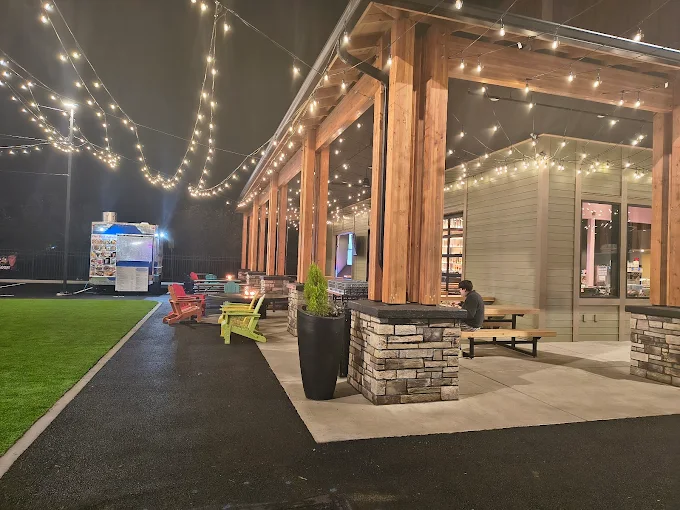 Outdoor patio area at night with string lights overhead, wooden columns, colorful Adirondack chairs, and a person sitting at a picnic table.