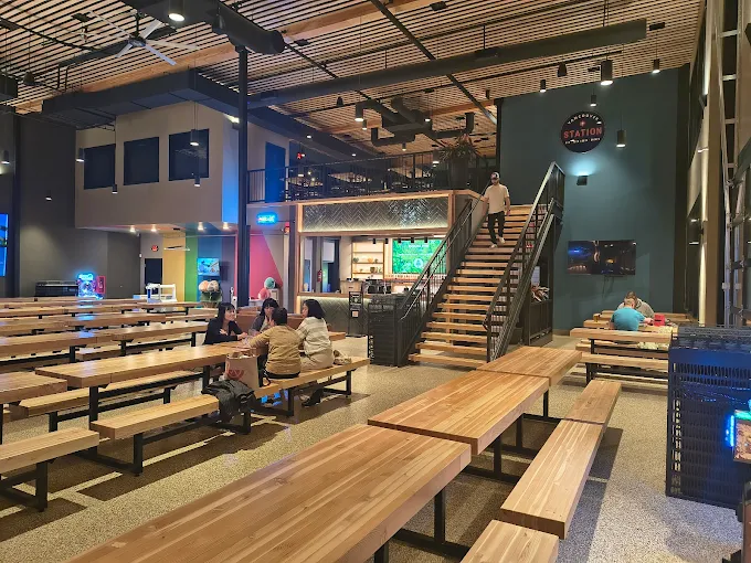 Modern indoor dining area with wooden picnic-style tables and benches, a staircase to an upper level, and several people seated and conversing.