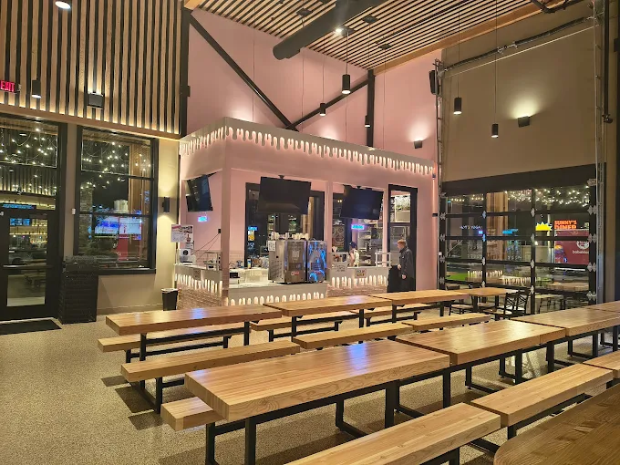 Indoor seating area with wooden picnic-style tables and benches facing a pink service counter decorated with white drip designs and multiple TV screens.