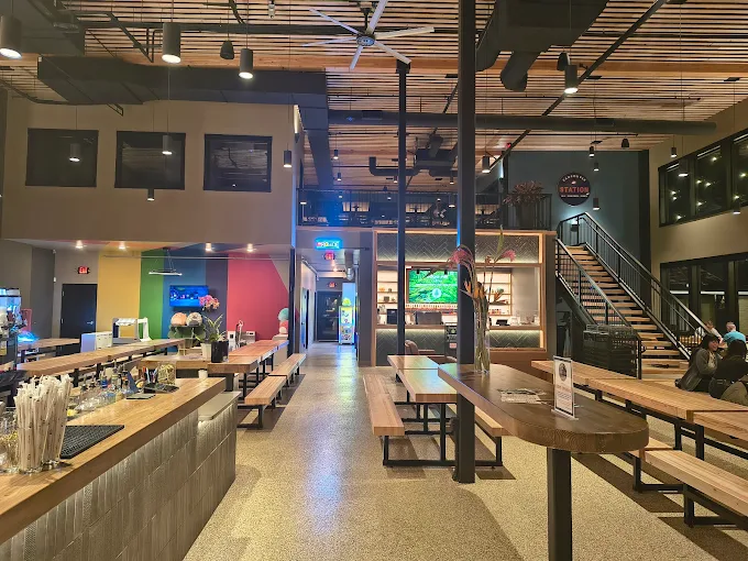 Modern indoor dining area with wooden picnic tables, a bar counter, stairs, ceiling fan, and colorful wall in the background.