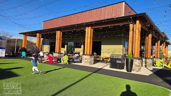 People sitting and a child running on artificial grass in front of a wooden pavilion with string lights on a sunny day.