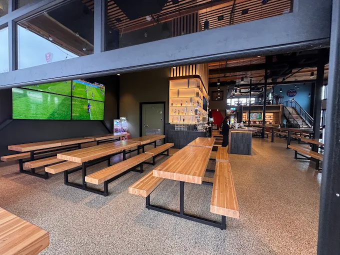 Modern sports bar interior with multiple wooden picnic-style tables and benches, large TV screens showing a soccer game, and a bar area with illuminated shelves.