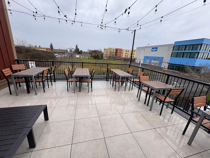 Outdoor patio area with empty tables and wooden chairs on tiled floor under string lights on a cloudy day.