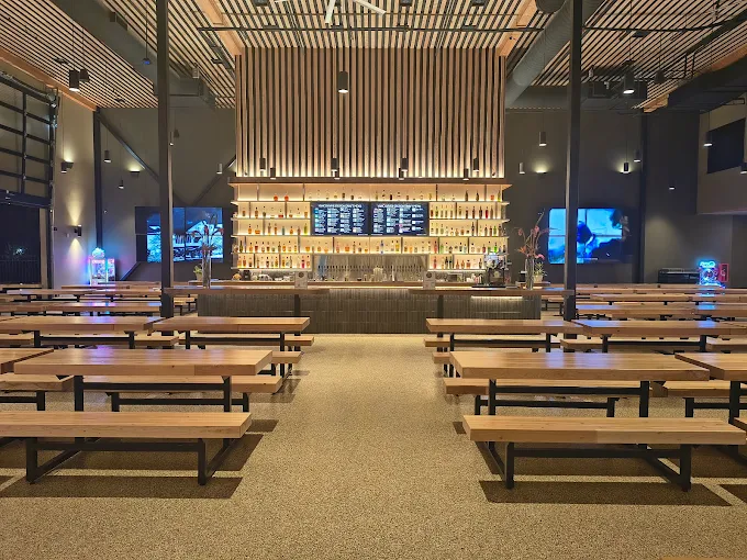 Modern bar interior with wooden benches and tables facing a well-lit bar counter stocked with bottles and digital menu screens.