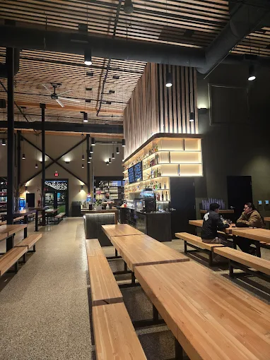 Interior of a modern café with wooden tables, benches, ceiling fans, and illuminated shelves behind the counter where two people are seated.
