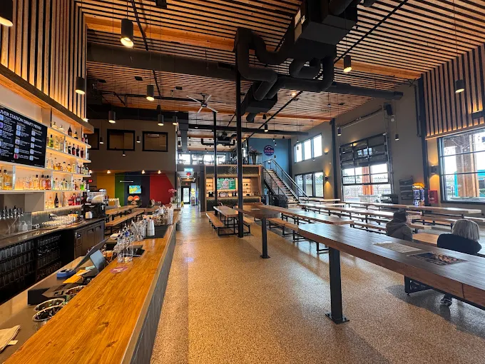Interior of a modern brewery taproom with long wooden tables, a wooden bar on the left, and industrial ceiling lighting.