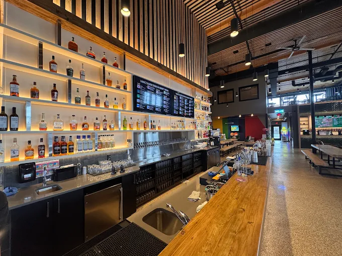 Modern bar interior with wooden countertop, shelves stocked with liquor bottles, and digital menu screens above the bar.