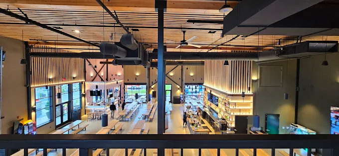 Spacious, modern restaurant interior with wooden tables, benches, and a well-lit bar area under a ceiling with wooden beams and industrial ductwork.