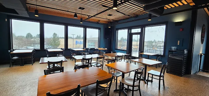 Empty restaurant dining area with wooden tables, black chairs, large windows, and a modern ceiling with hanging lights.