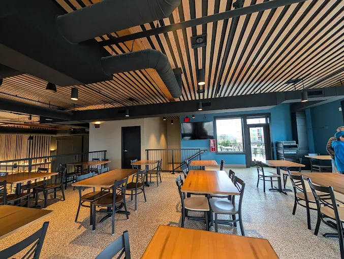 Modern dining area with wooden tables and chairs under a ceiling with black ventilation ducts and wooden slats.