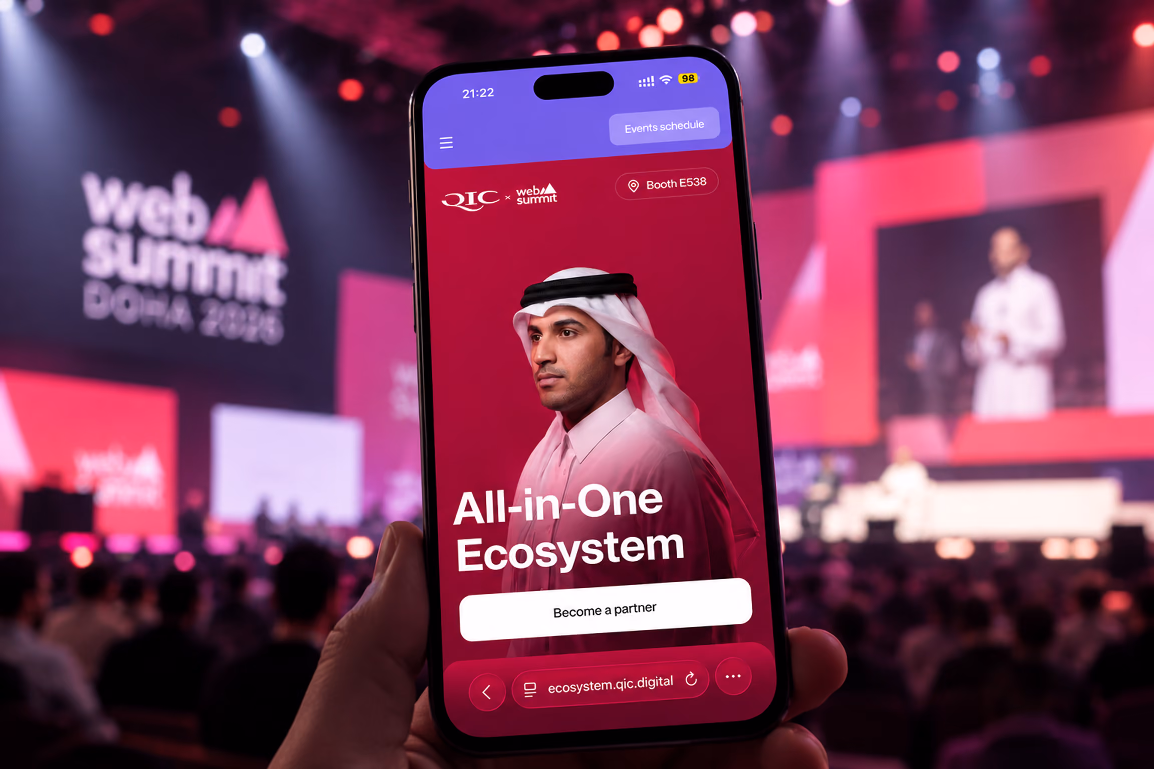 A hand holding a smartphone showing a website with a man in traditional Middle Eastern attire and text that reads 'All-in-One Ecosystem' and 'Become a partner' at a crowded Web Summit event with blurred stage and audience in the background.