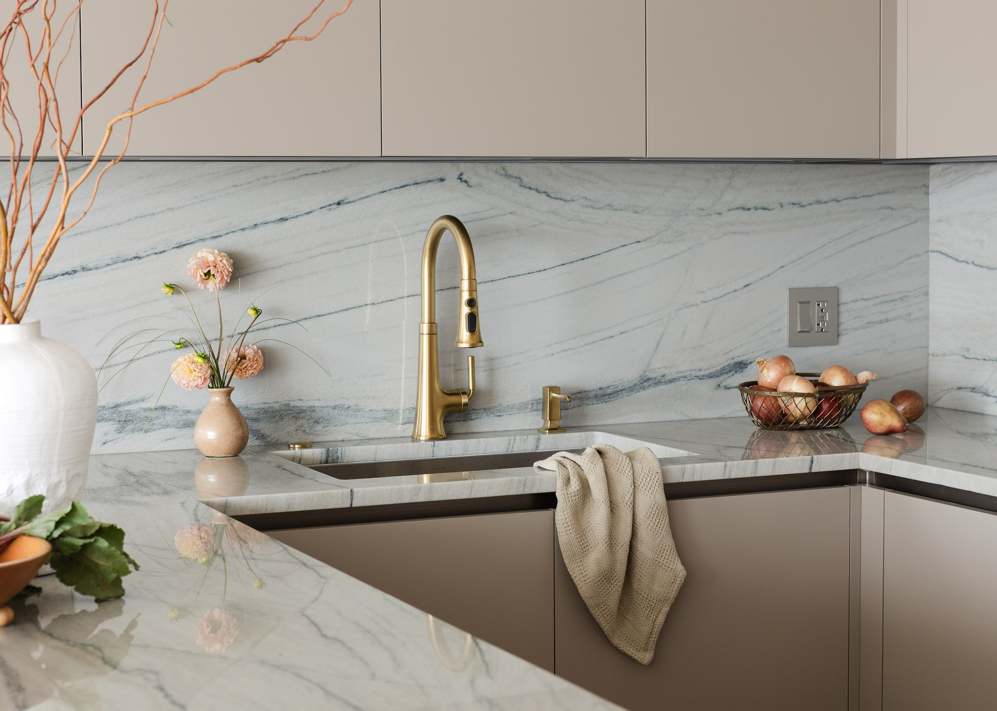 A kitchen tap in marble and beige kitchen. The kitchen tap is a gold color. On the left side of the sink is a vase with flowers inside and on the right is a basket of onions.