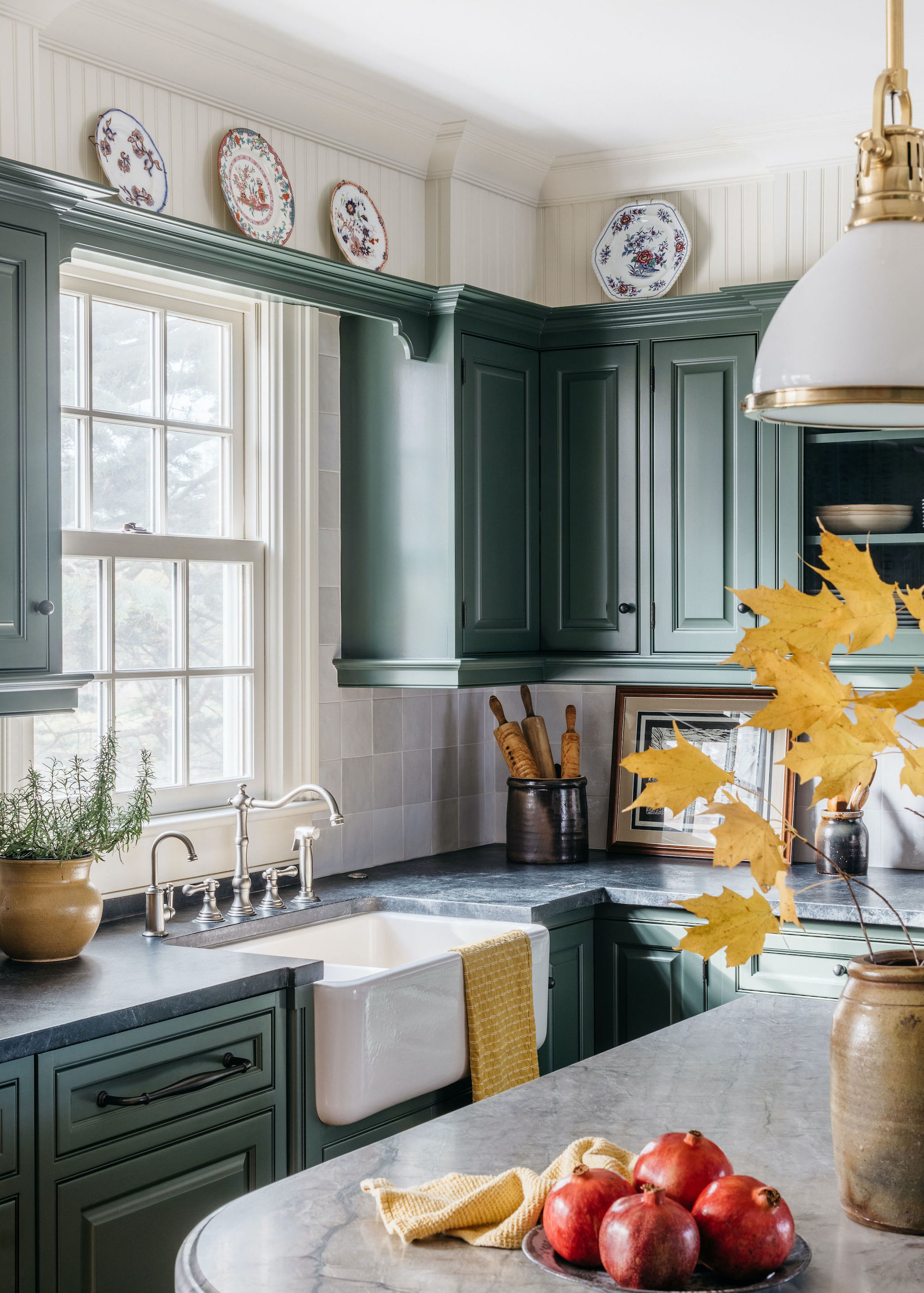 A kitchen tap in a green kitchen with a belfast sink. Beside the sink is a plant in a large vase/pot. There is also a kitchen island with pomegranates on top.