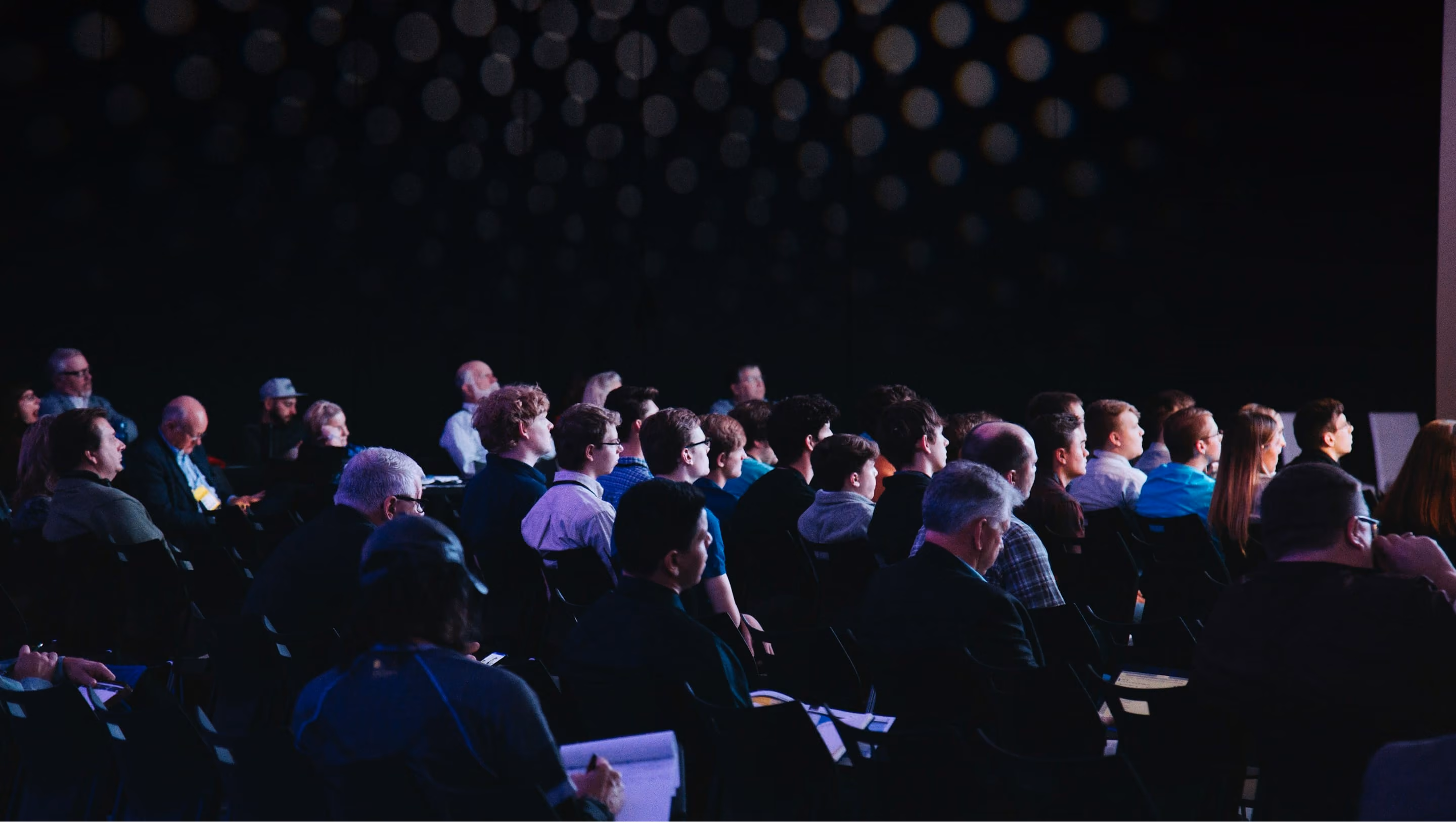 Audience seated in rows facing a stage in a dimly lit conference or event room.