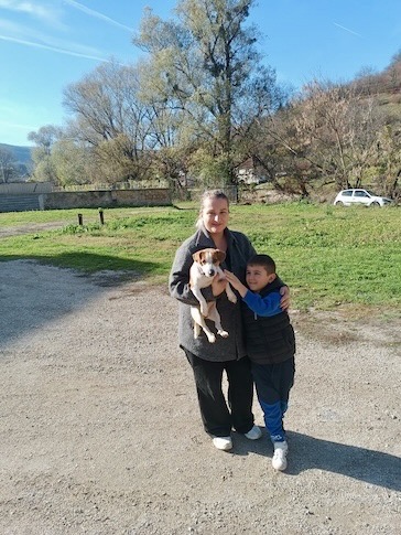 Eine Frau hält einen kleinen Hund, während ein Junge sie umarmt, im Freien auf einem sonnigen Weg mit Bäumen und einem Auto im Hintergrund.