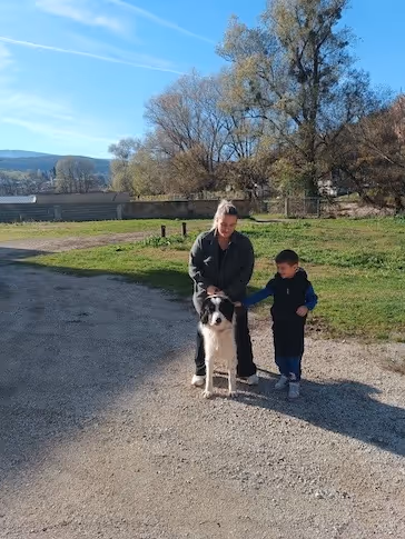 Frau und Junge stehen zusammen auf einem Feldweg neben einem großen schwarz-weissen Hund, sonniger Tag mit Bäumen und Wiese im Hintergrund.