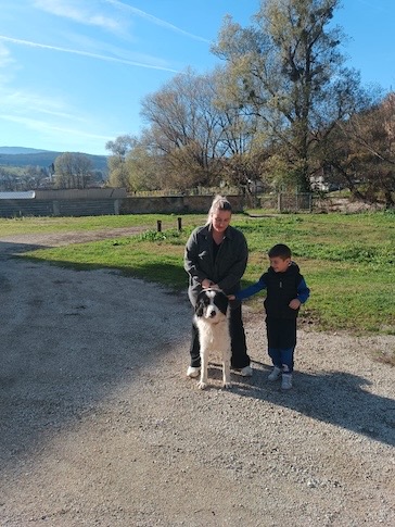 Frau und Junge stehen zusammen auf einem Feldweg neben einem großen schwarz-weissen Hund, sonniger Tag mit Bäumen und Wiese im Hintergrund.