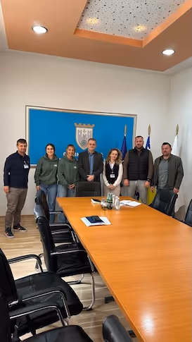 Sieben Personen stehen in einem Sitzungsraum vor einer blauen Wand mit einem Wappen, daneben sind mehrere Flaggen aufgestellt.