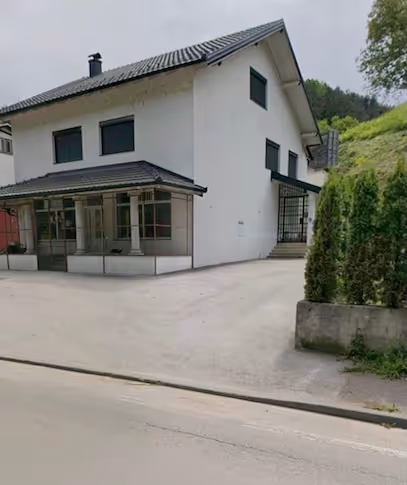 Weisses, zweistöckiges Haus mit schwarzem Dach an einer leeren Strasse, umgeben von Grünflächen und Büschen.