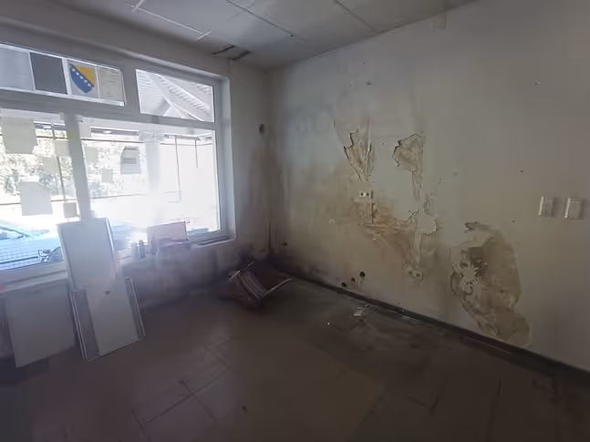 Verlassener Raum mit abgeblätterten Wänden, umgekipptem Stuhl und großem Fenster mit Blick auf die Straße.