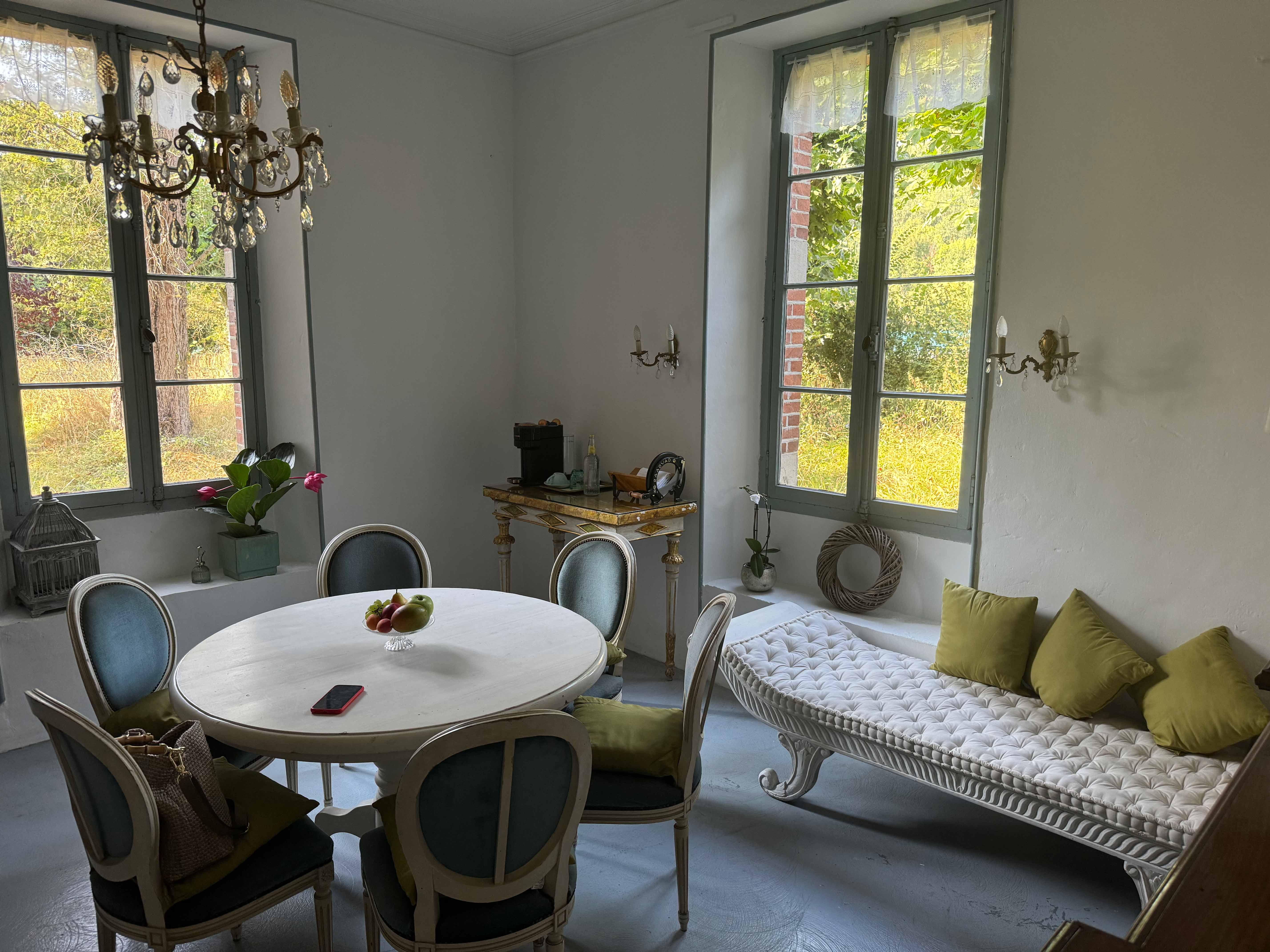 Elegantes Esszimmer mit runden weißen Tisch, sechs blaugrauen Stühlen, großem Fenster, Kamin und Kronleuchter.