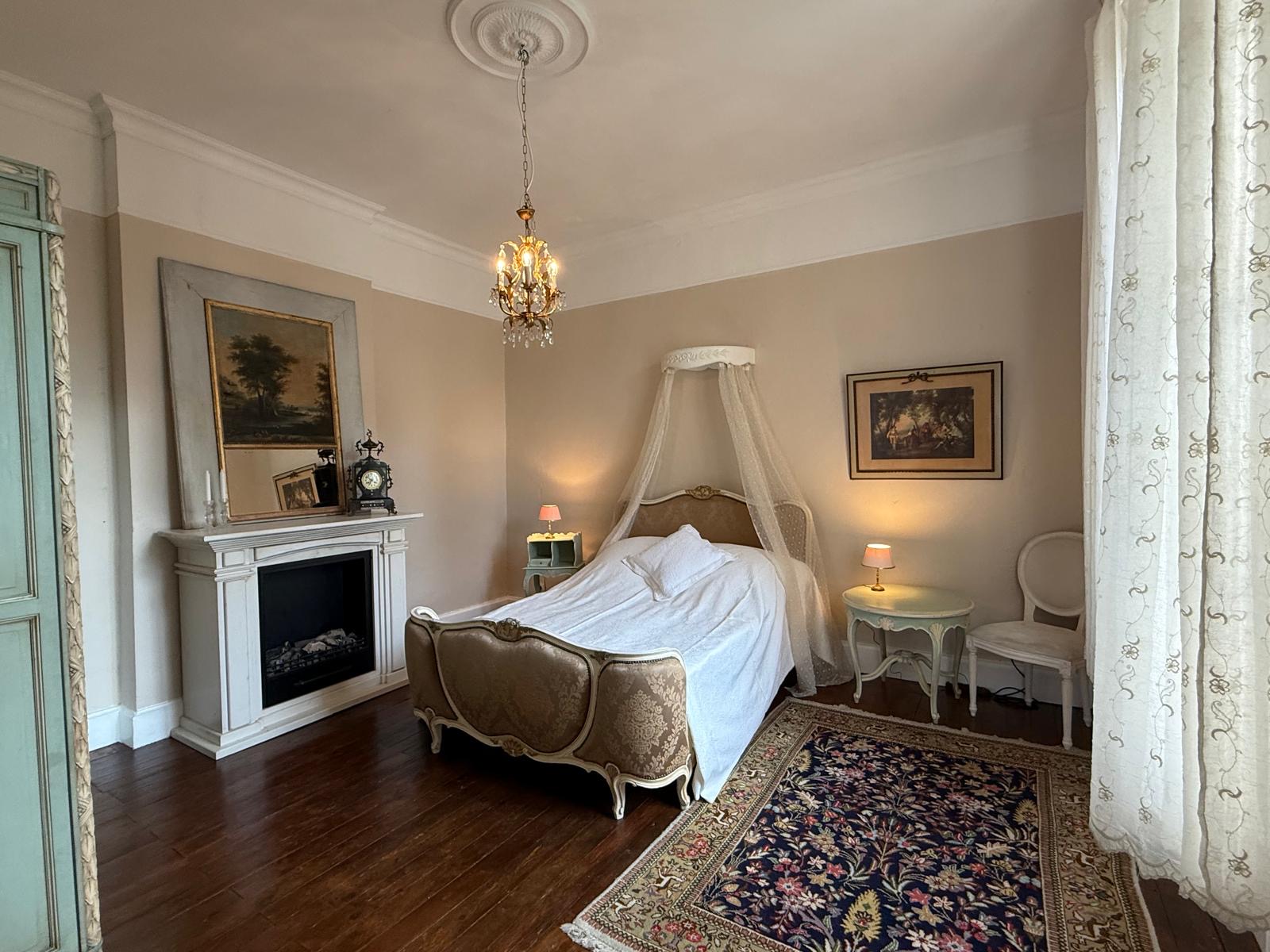 Elegantes Schlafzimmer mit einem Bett mit Baldachin, antikem Kamin, Kronleuchter und gemustertem Teppichboden.