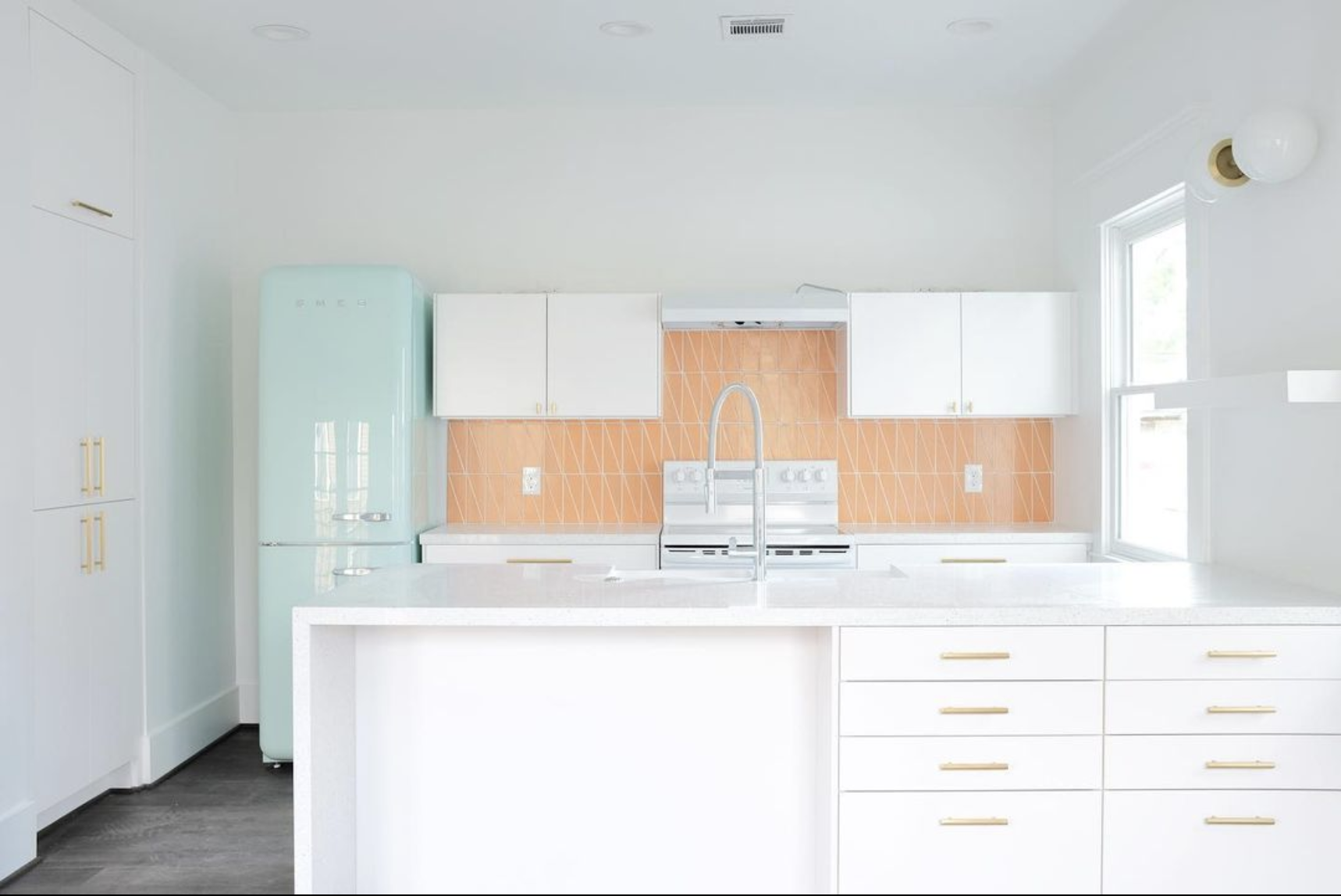 This kitchen features a salmon orange tile backdrop among beautiful white cabinets and fixtures. A retro, light aqua fridge frames the kitchen on the left.
