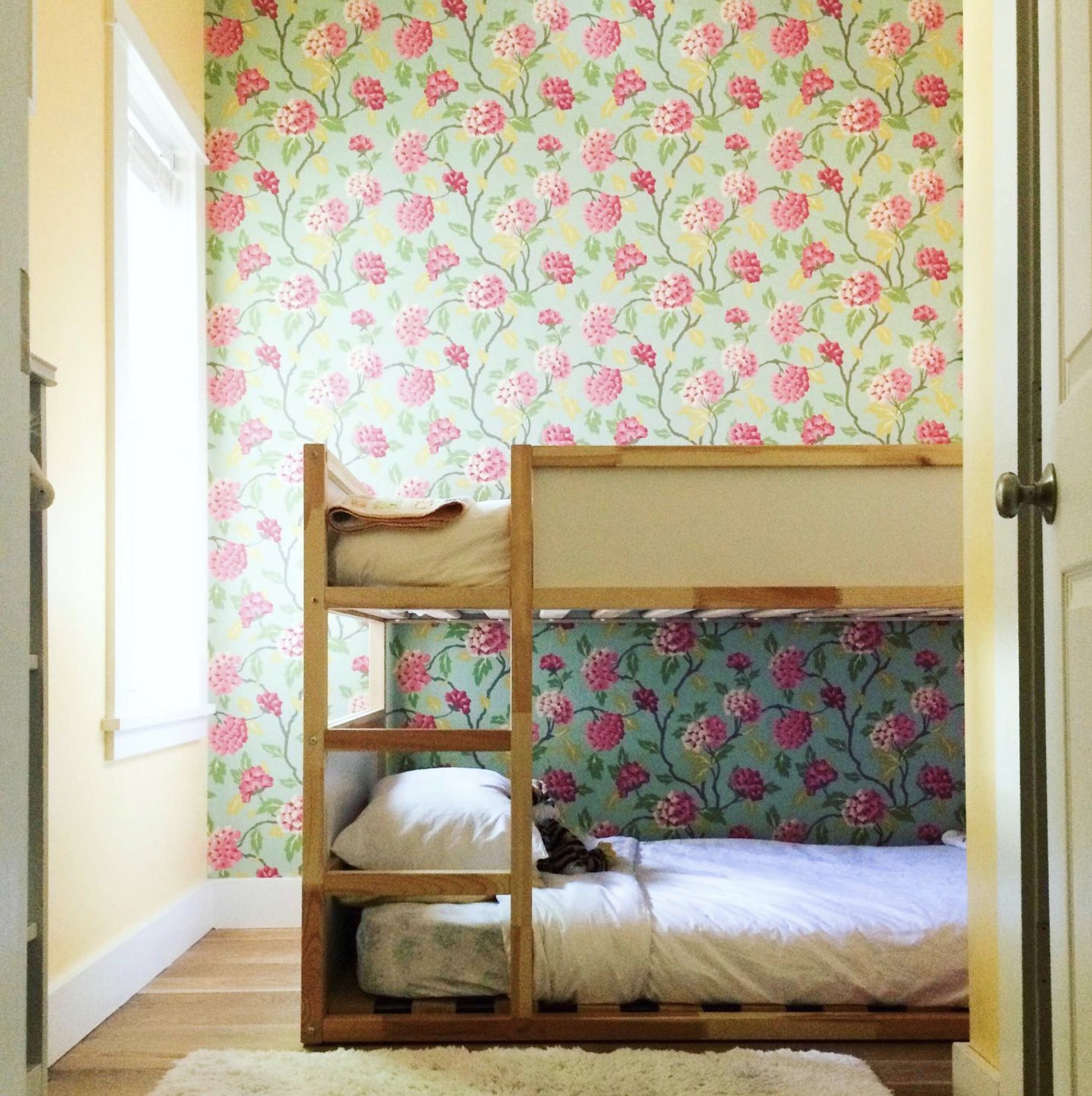 A two toned pink rose against a very light green background patterns the wallpaper behind a bunkbed for a child's room. The well lit area feels warm and cozy.