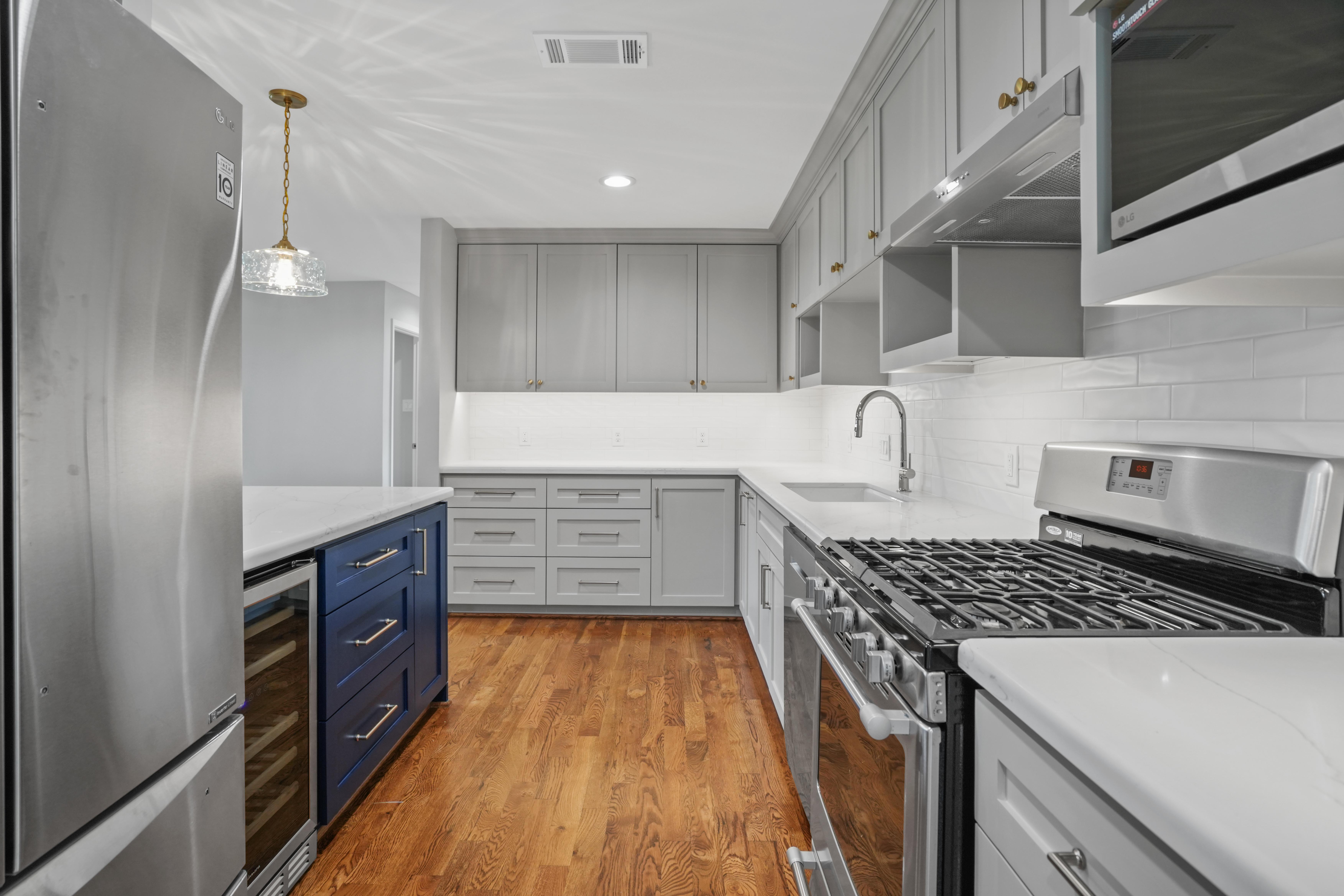This monochromatic renovated kitchen features grey cabinetry among white backsplash tiling. A pop of color shows behind the kitchen counter on some navy blue drawers.