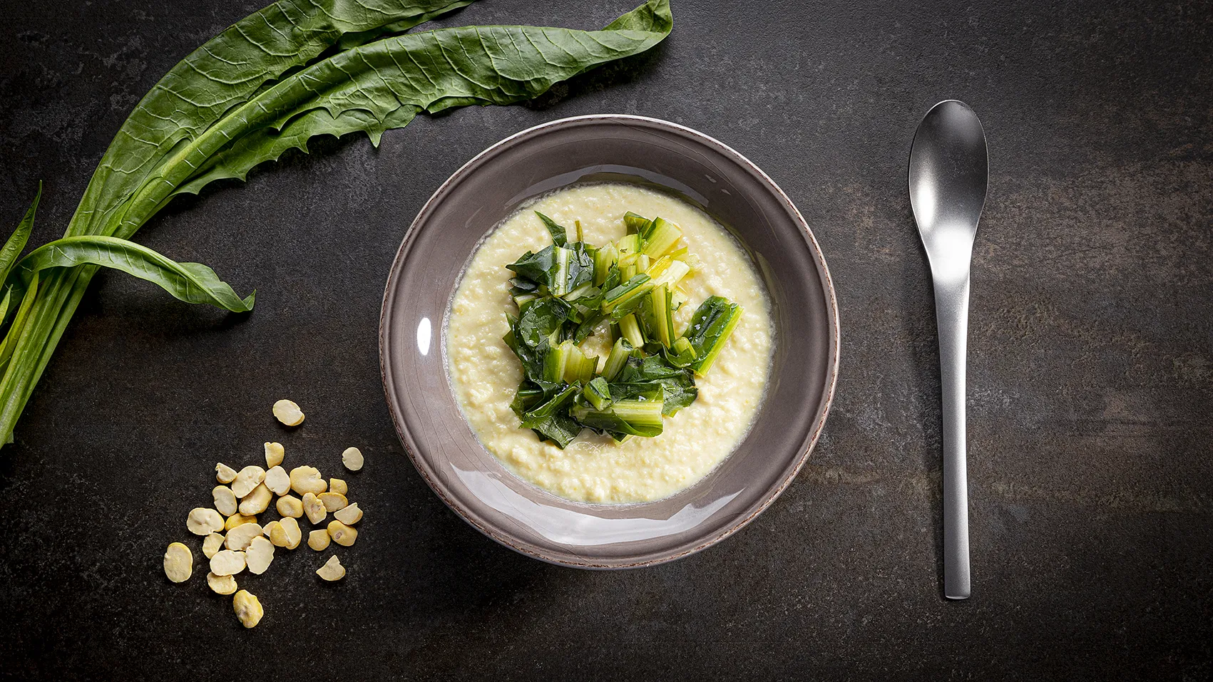 Ciotola di crema di polenta con bietole verdi al centro, accanto a foglie fresche e chicchi di mais secchi su superficie scura con un cucchiaio in acciaio.