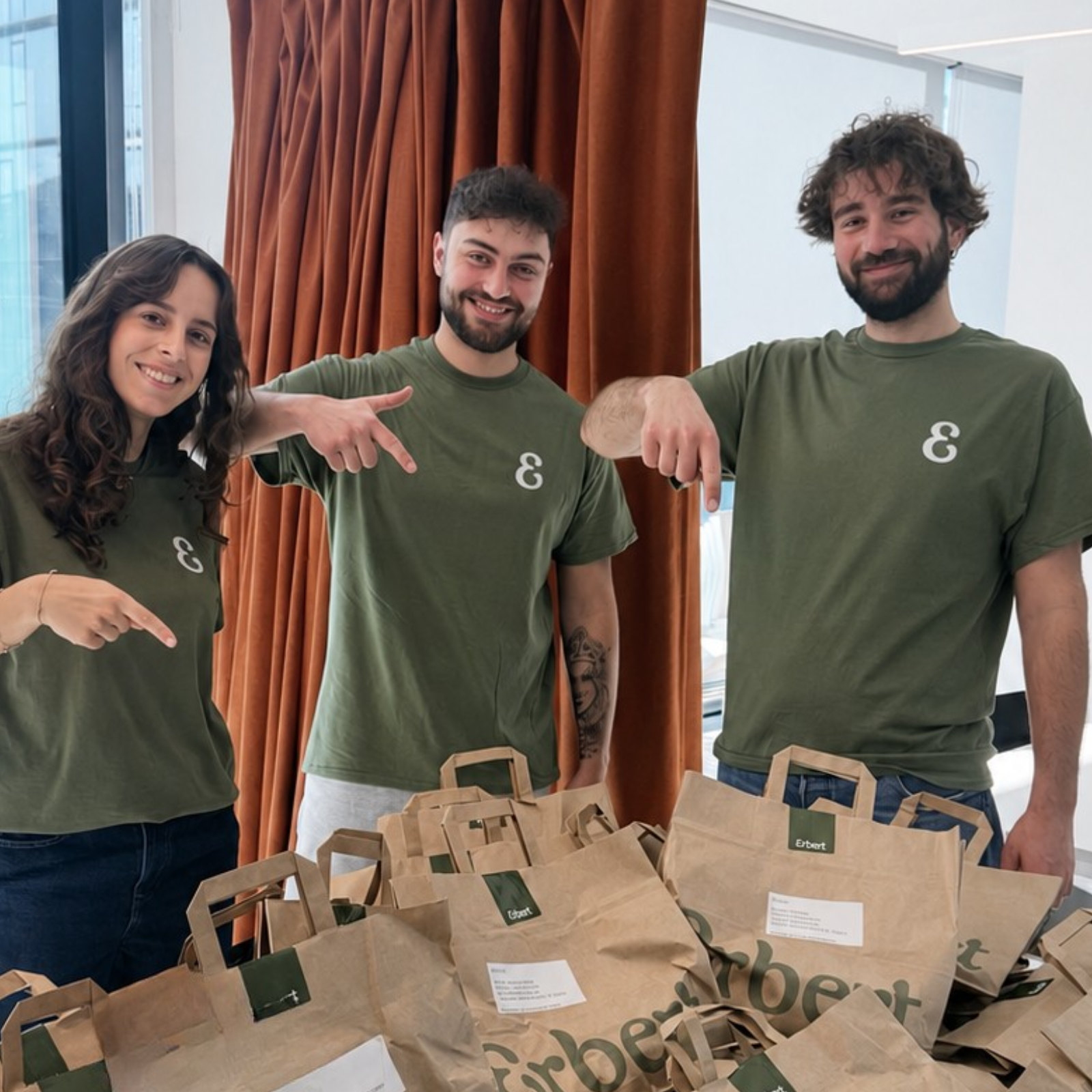 Uomo con maglione beige che siede su un divano grigio decorato per Natale, mangia un'insalata da un contenitore Erbert e tiene un bicchiere di carta.
