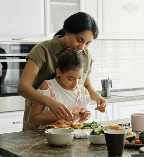 Woman and young girl preparing food together in a kitchen, placing leafy greens on bread slices at a counter.