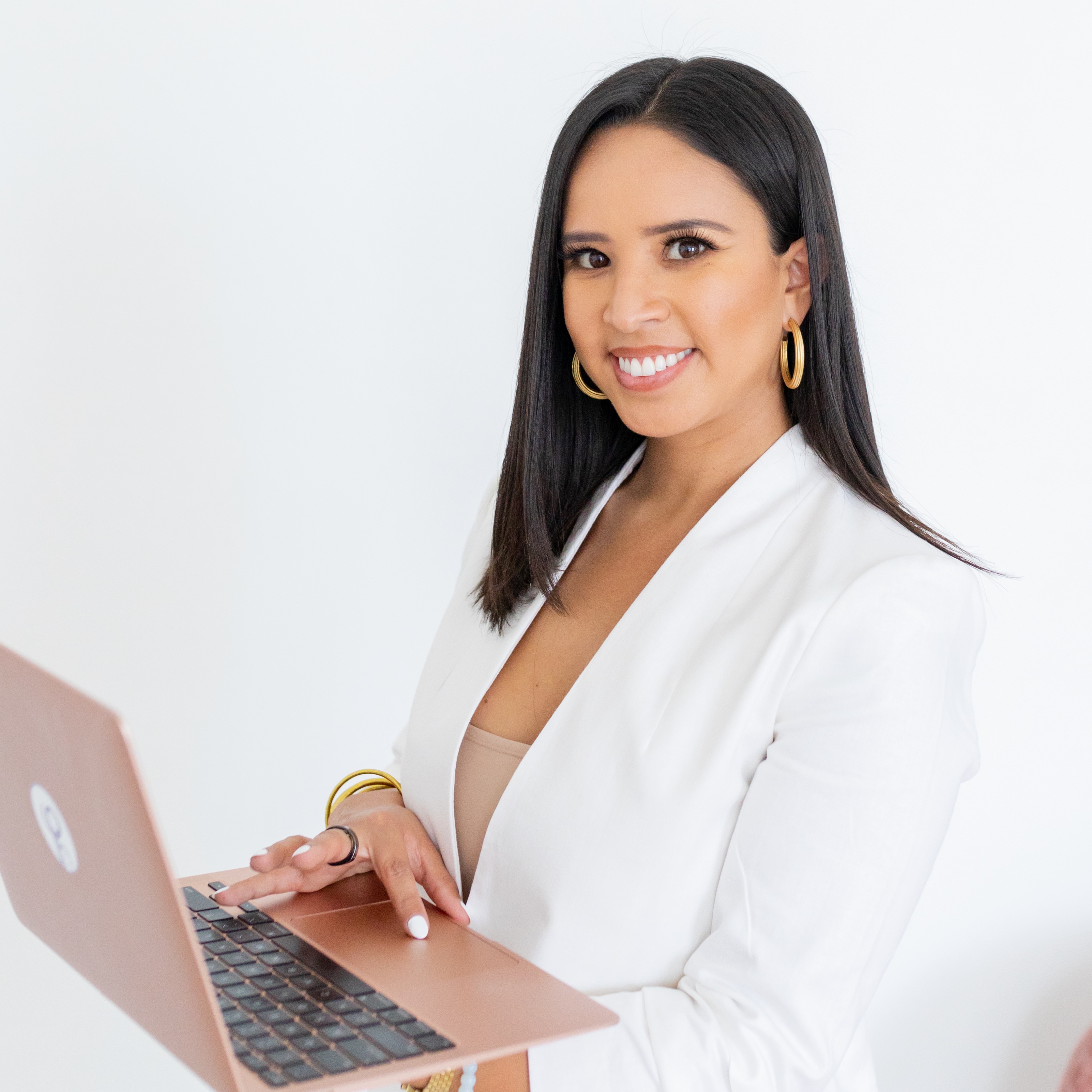 Smiling woman with long dark hair in a white blazer holding a rose gold laptop against a white background.