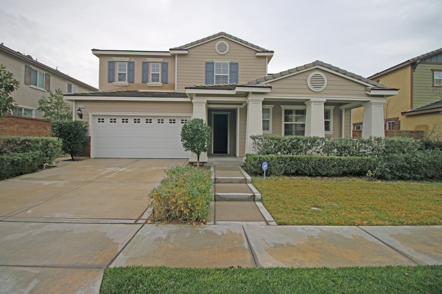 Two-story beige suburban house with blue shutters, white garage door, front porch, and manicured lawn with shrubs.