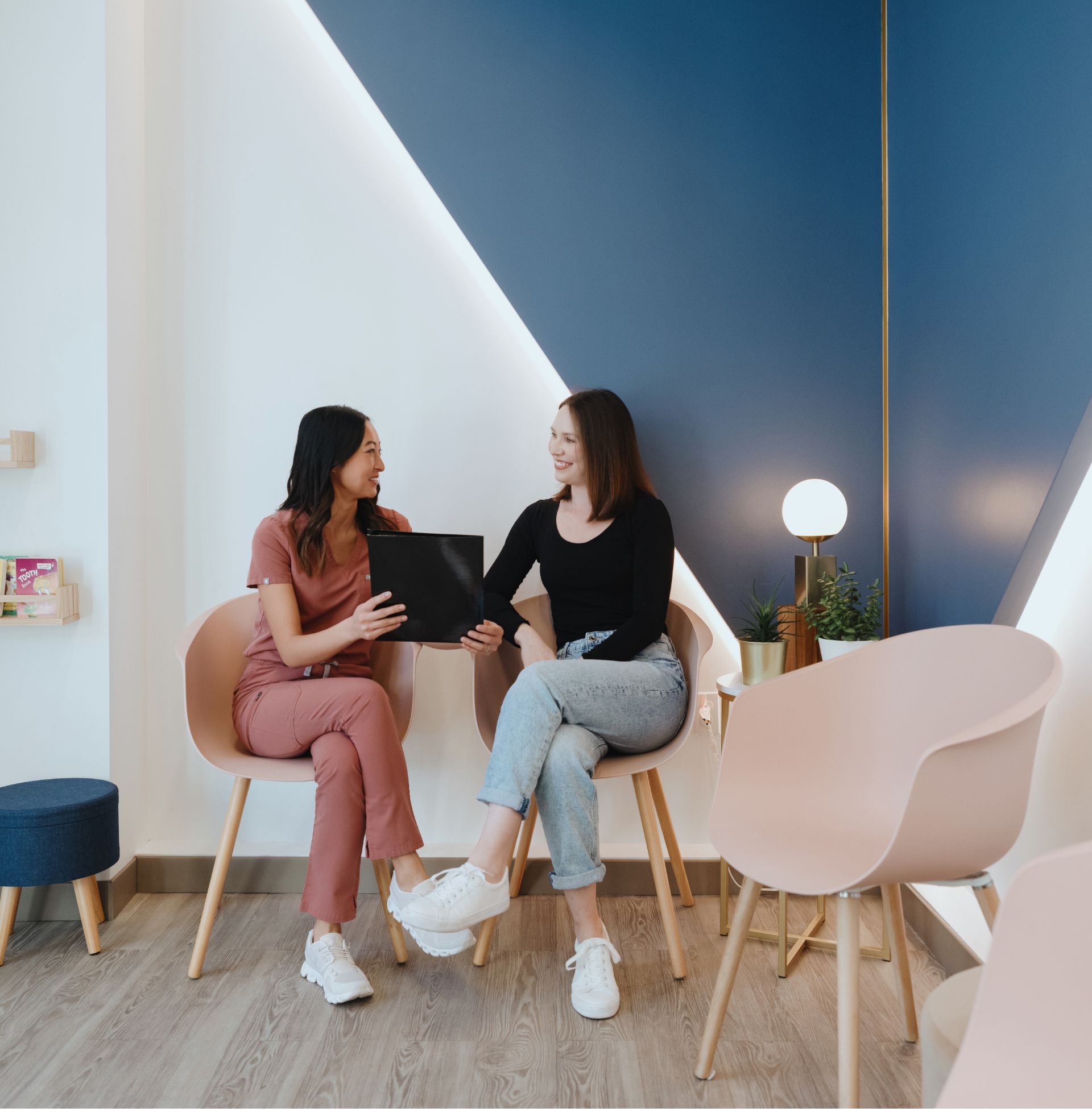 A healthcare professional in scrubs showing a black folder to a smiling woman sitting in a modern waiting room.