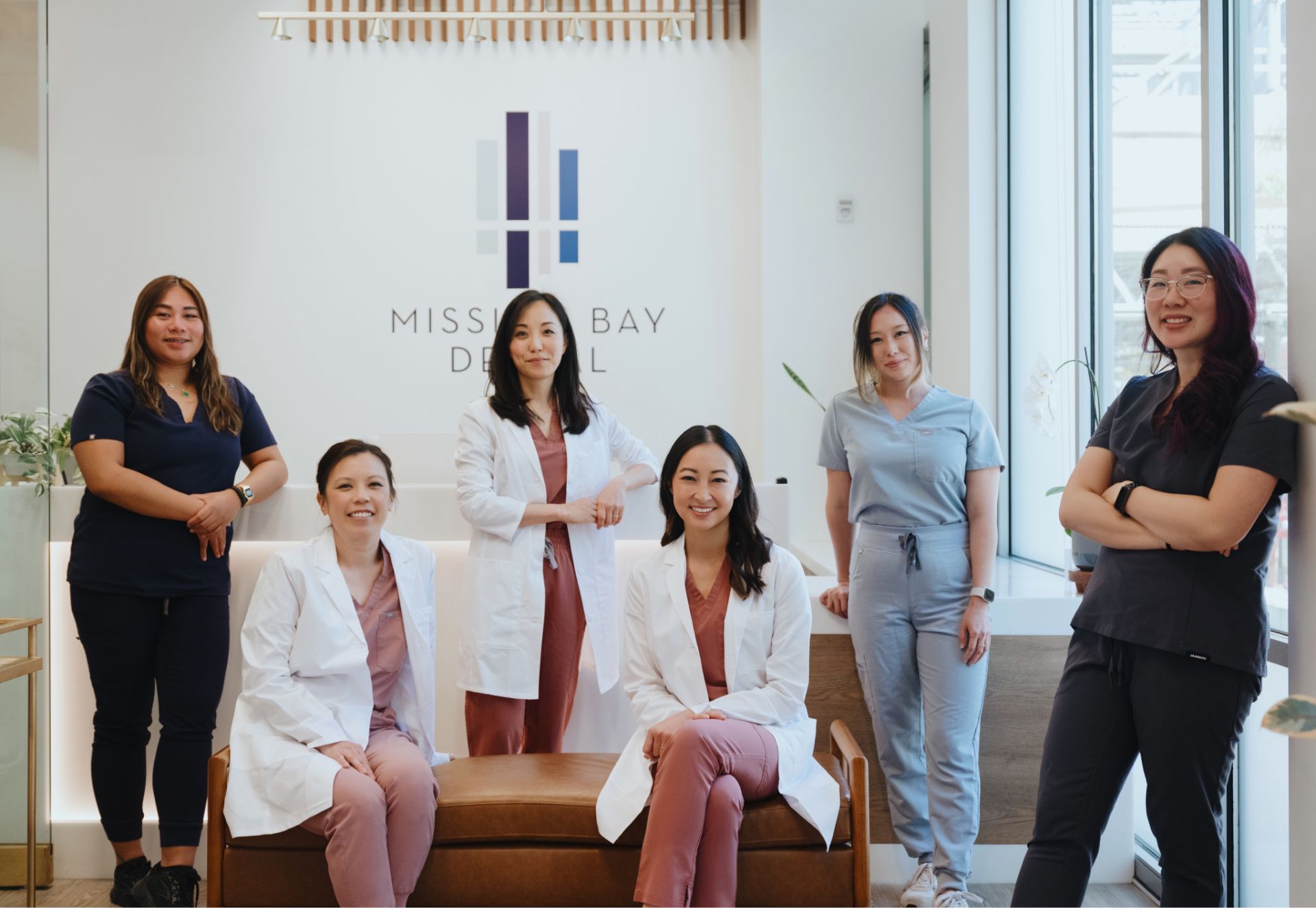 Six female dental professionals posing inside Mission Bay Dental office, dressed in scrubs and lab coats.