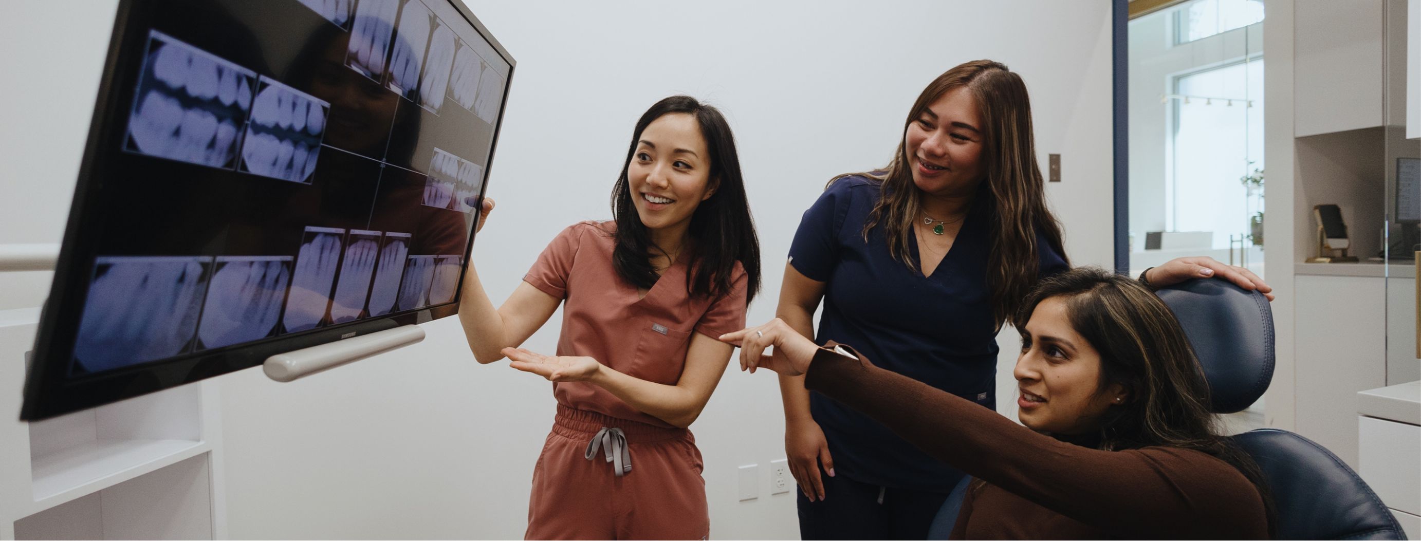 A dental professional shows dental X-rays on a monitor to two women, one seated in a dental chair and pointing at the images.
