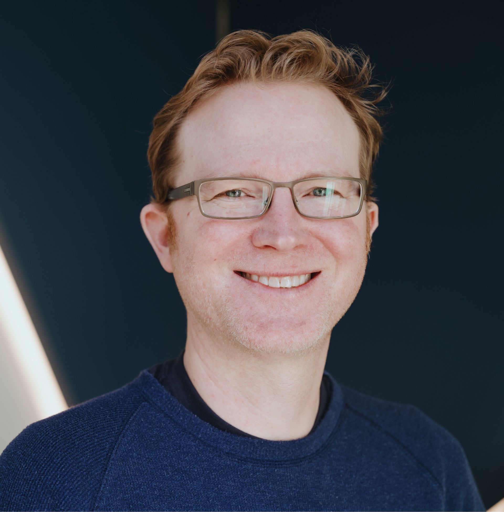 Smiling man with glasses, short light brown hair, and a navy blue sweater against a dark background.