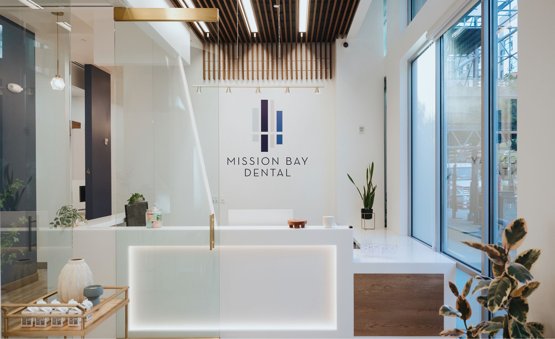 Modern dental office reception area with illuminated white desk, large windows, plants, and Mission Bay Dental logo on the wall.