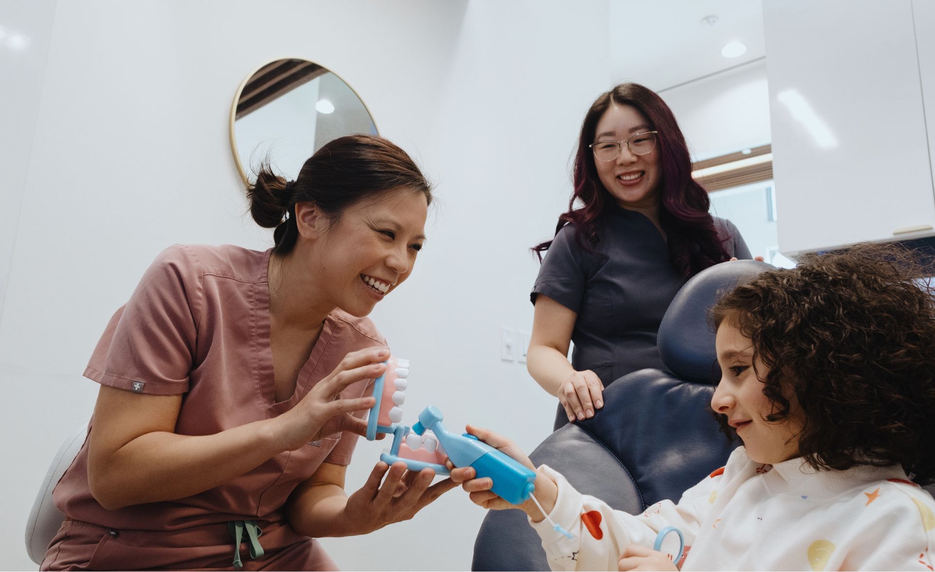 Female dental professional smiling and demonstrating teeth cleaning using a dental model and toothbrush with a young girl in a dental chair, while another woman watches.