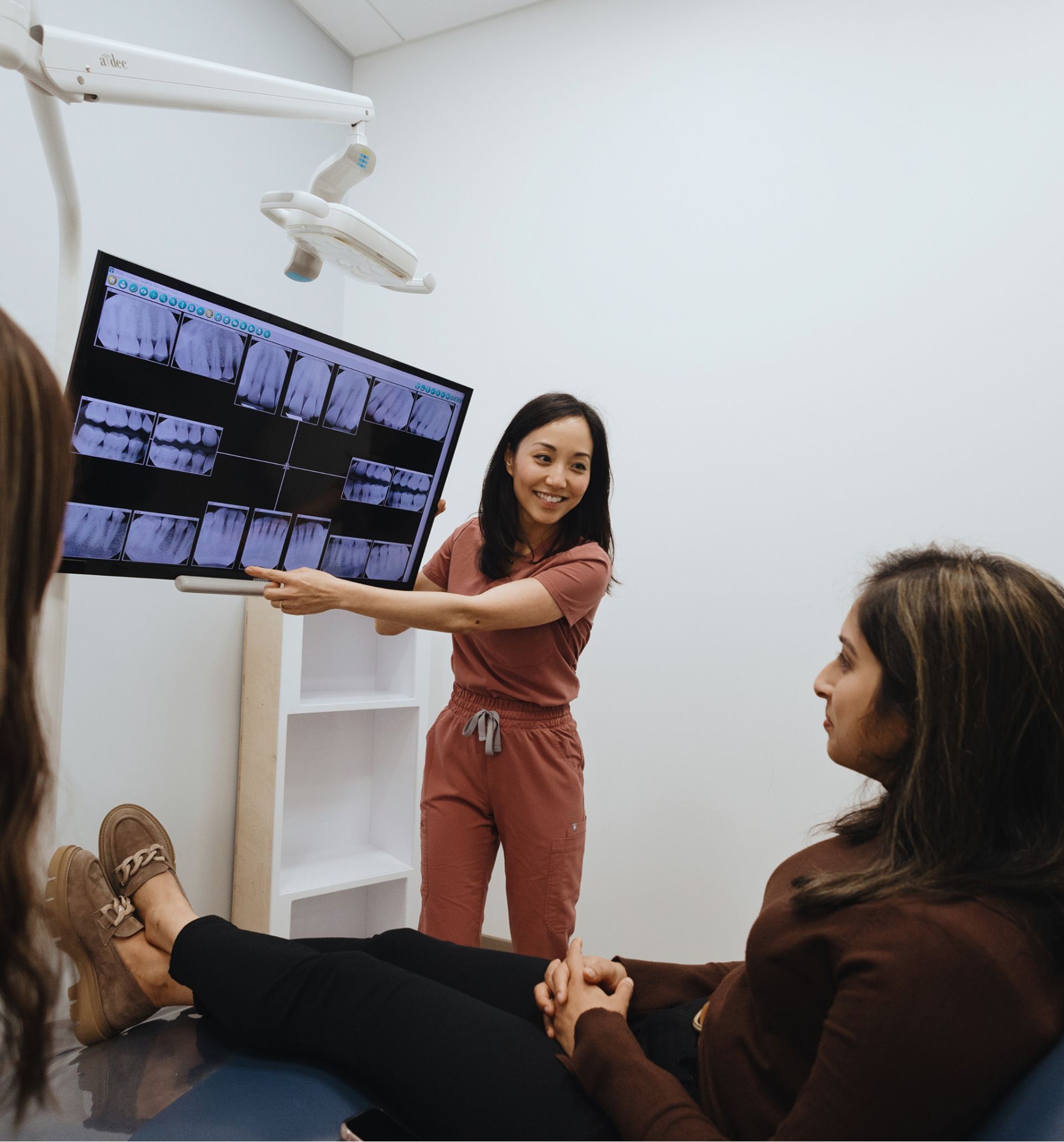 Smiling female dentist pointing at dental X-rays on a screen while talking to a female patient seated on a dental chair.
