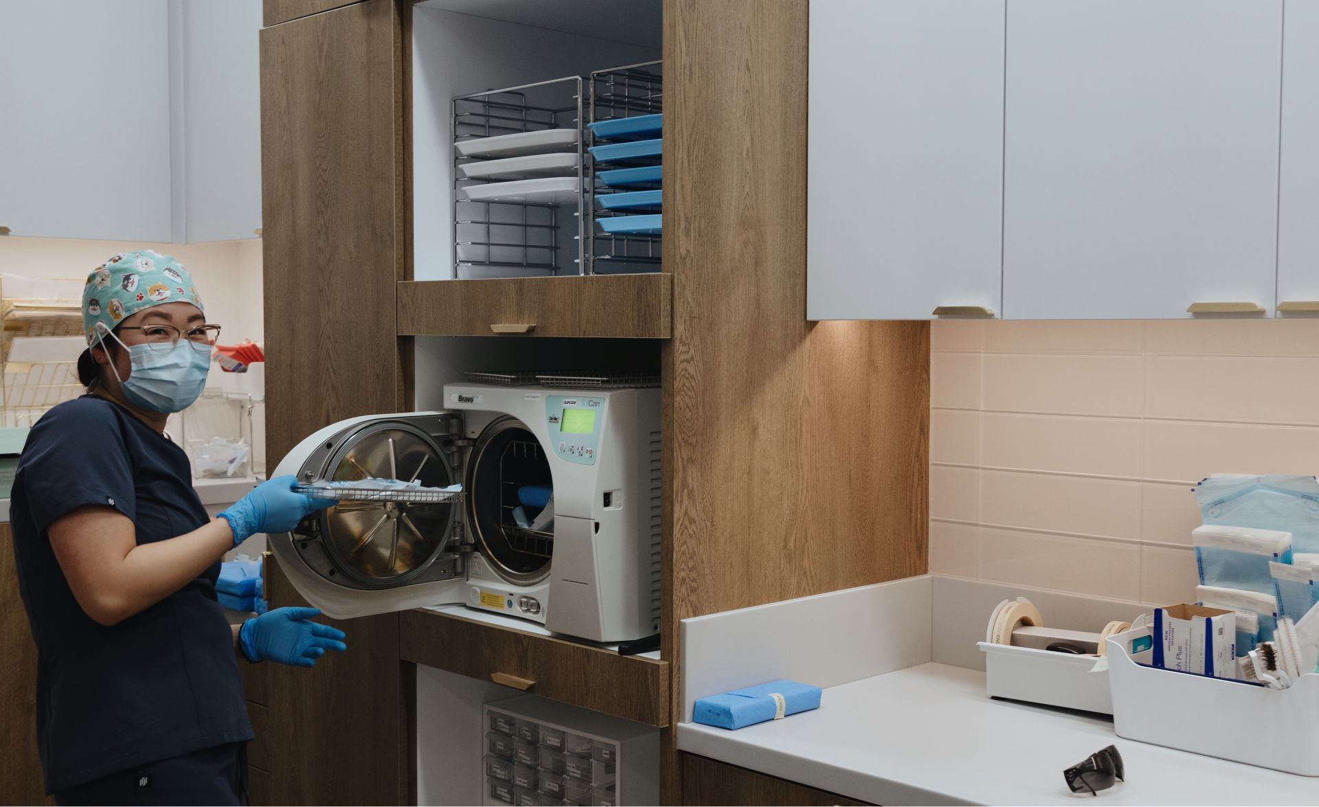 Healthcare worker in scrubs, gloves, mask, and cap operating a sterilization machine in a medical supply room.