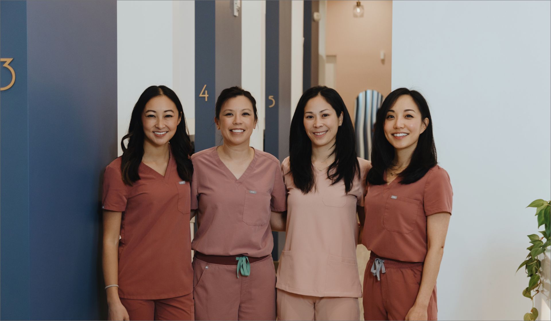 Four smiling female medical professionals wearing scrubs standing together in a clinical corridor.