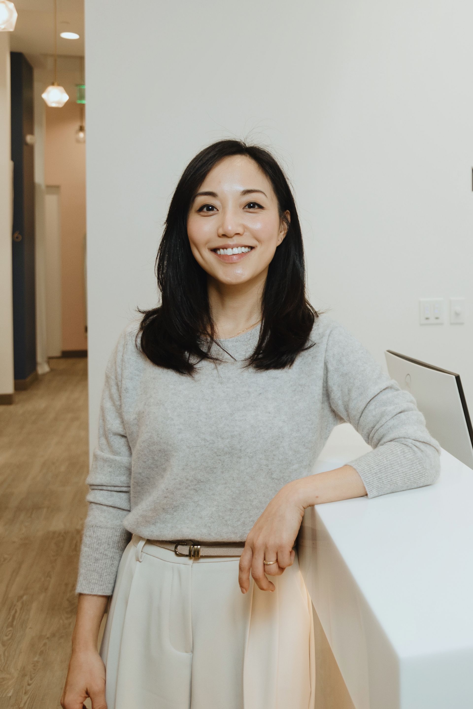 Smiling woman with black hair wearing a light grey sweater and beige pants leaning on a white counter.