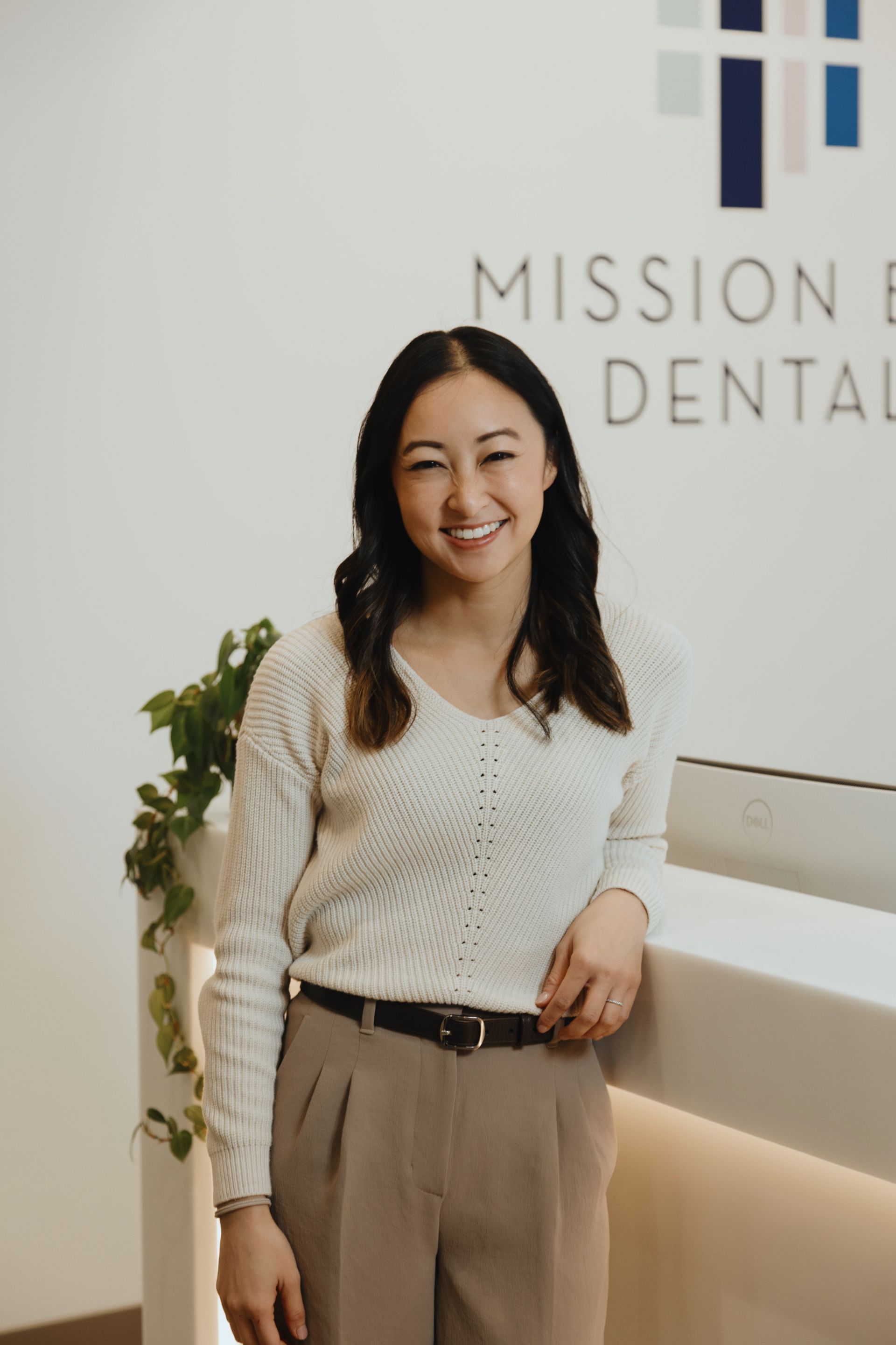 Smiling woman in a white sweater and beige pants standing near a dental office reception desk with a plant in the background.